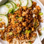 Crumbled tempeh on brown rice with cucumber slices in a white bowl.