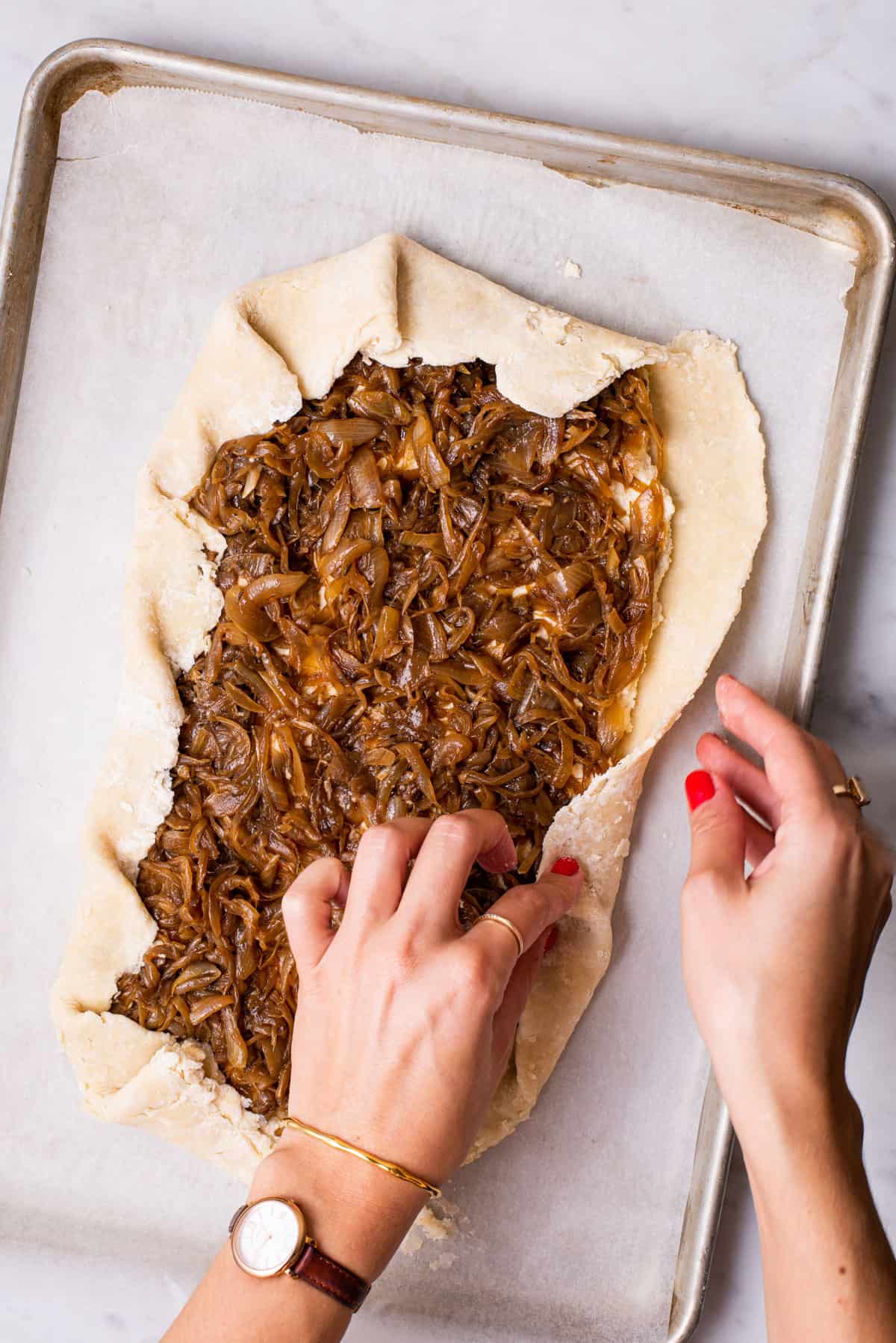 Woman's hands folding border of unbaked caramelized onion galette on a baking sheet.