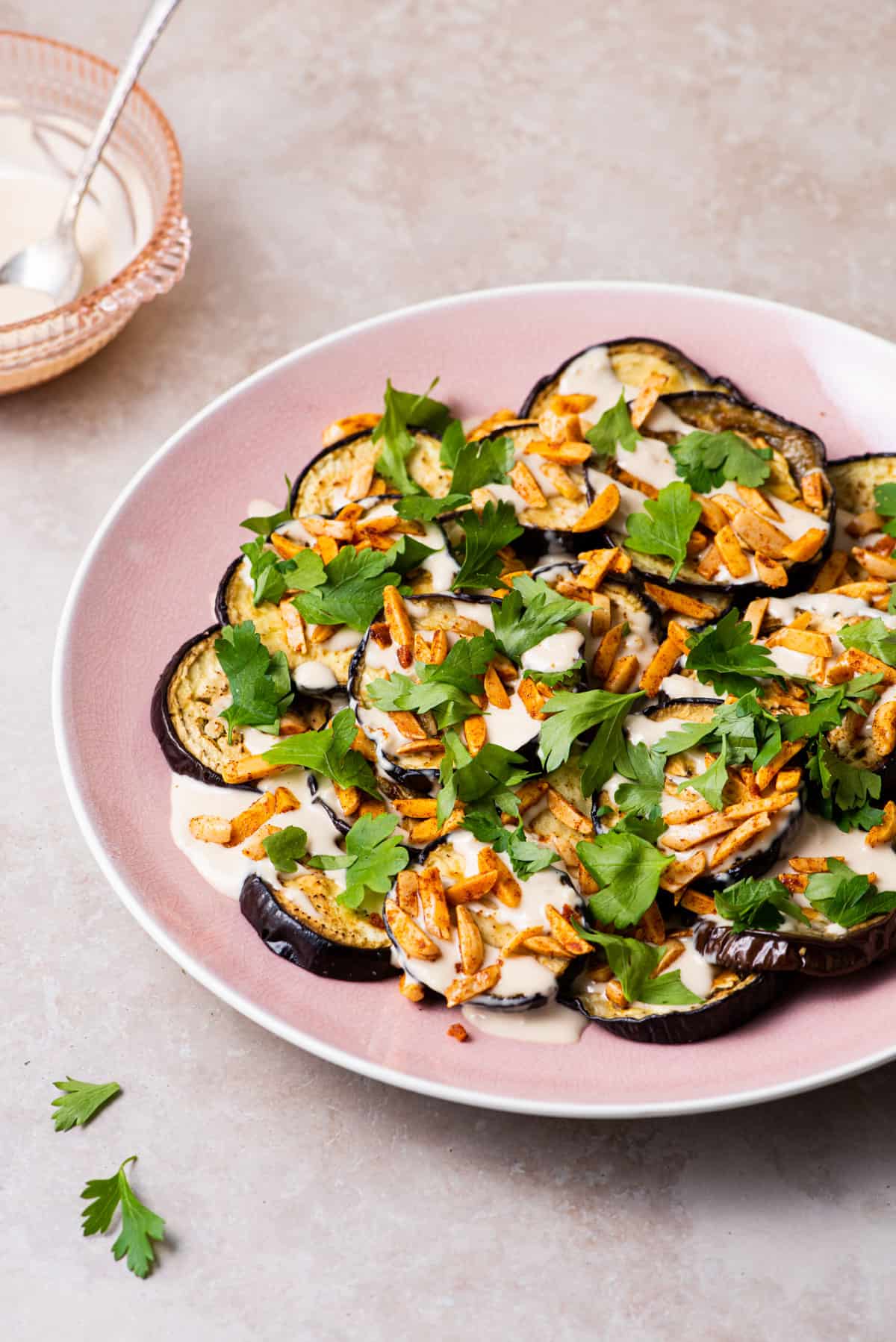 Ottolenghi inspired roasted eggplant rounds on a pink platter, drizzled with tahini sauce and topped with spiced almonds and parsley.