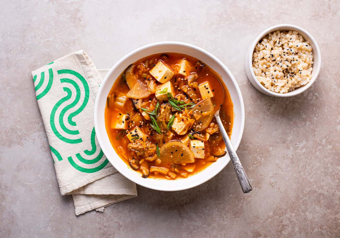 Tofu kimchi stew with daikon in a white bowl next to scallions and sesame seeds.