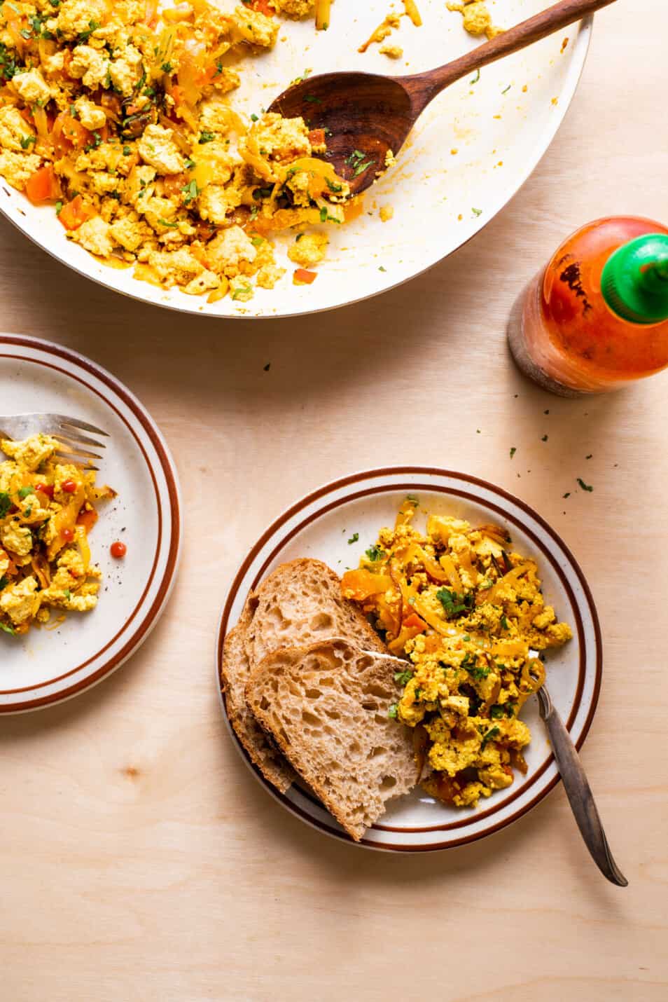 Vegan brunch recipe for tofu scramble with onions and tomatoes on a plate with toast next to sriracha.