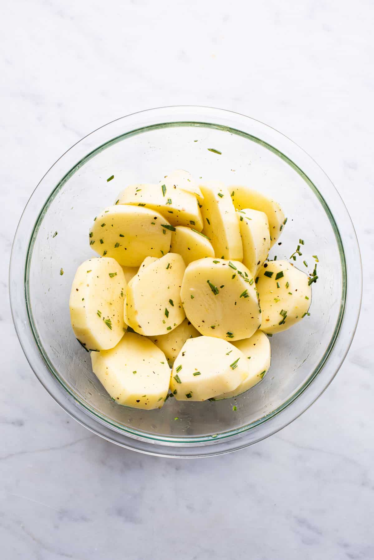 Sliced potatoes in a glass bowl with oil and rosemary.