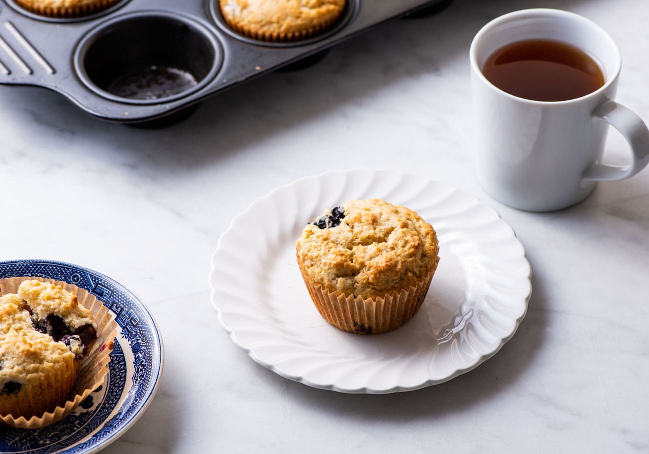 Vegan blueberry lemon muffin on a white plate next to a cup of tea.