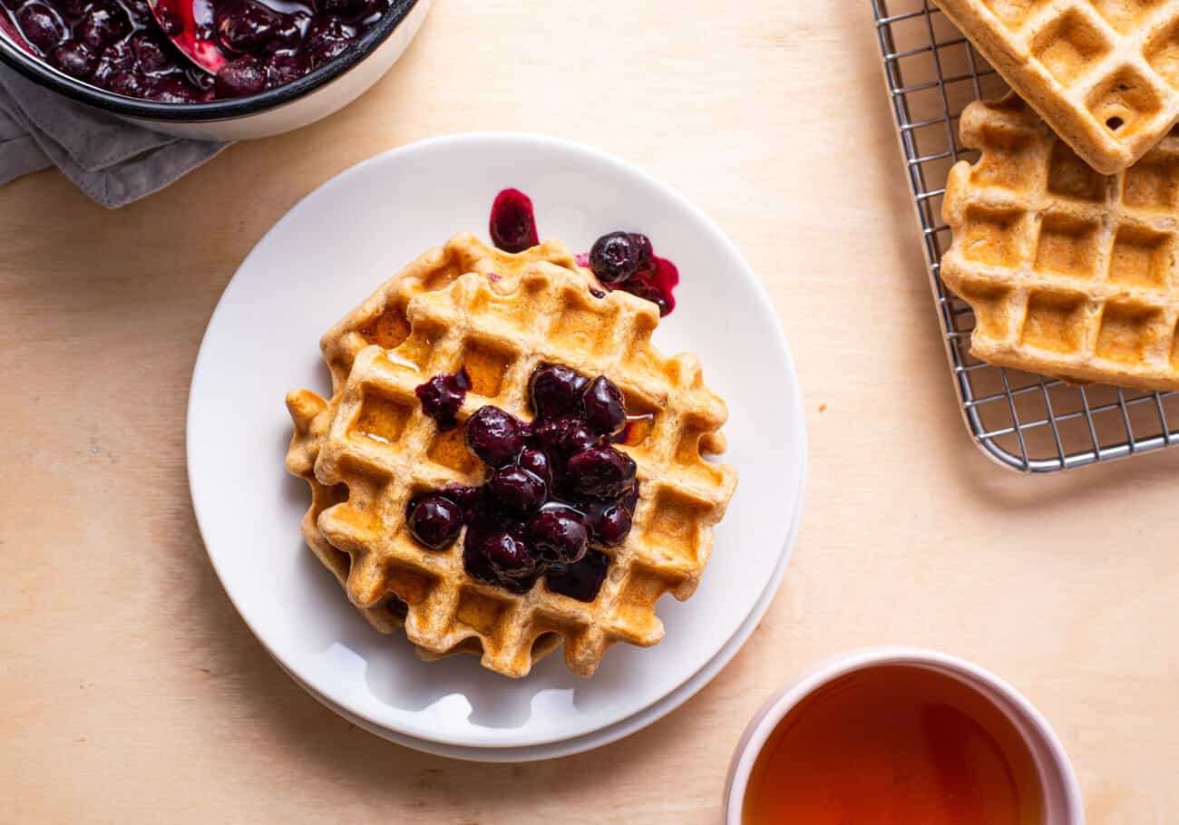 Oat waffles on a white plate with blueberry compote next to a tea cup.