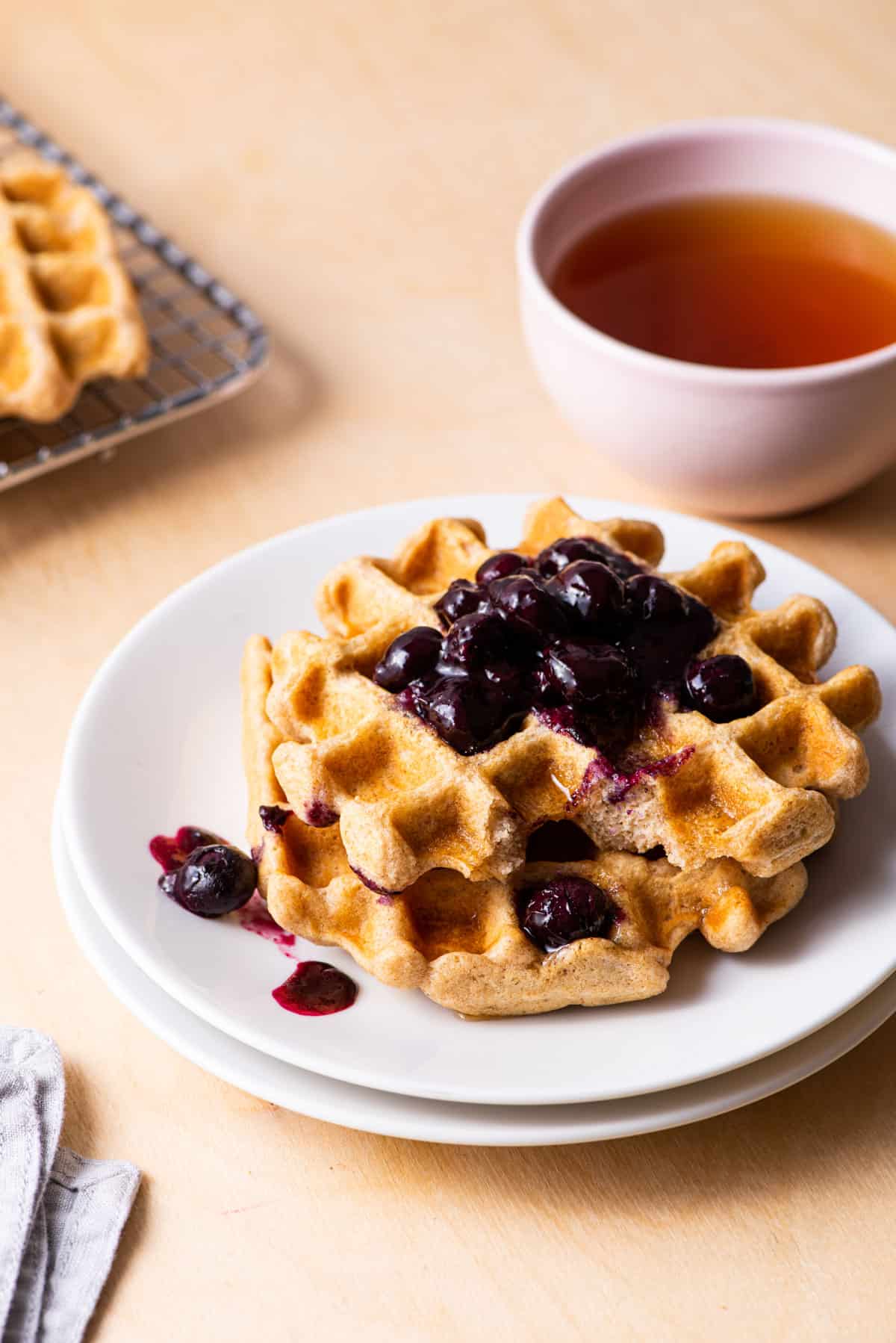 Vegan oat waffles on a white plate with blueberry compote, next to teacup.