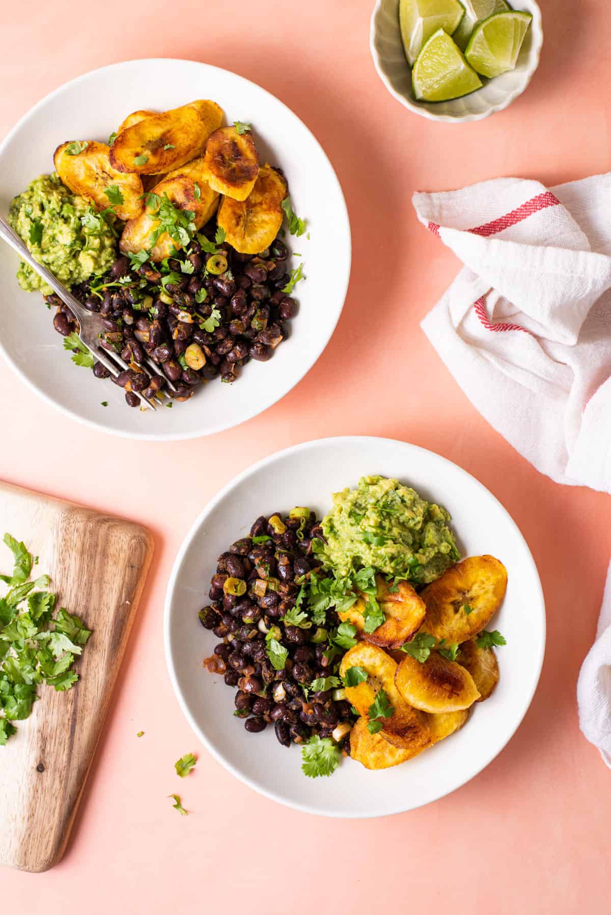 Black beans and plantains in white bowls with guacamole, next to cilantro and limes.