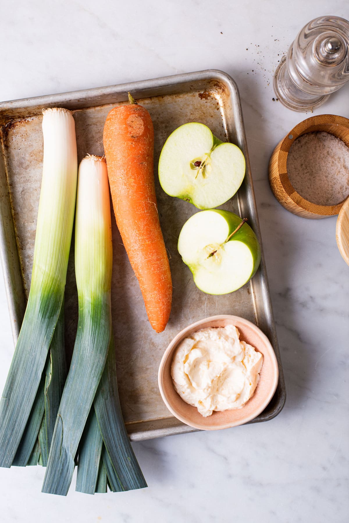 Leeks, a carrot, a green apple, mayo, salt, and pepper gathered on a marble table.