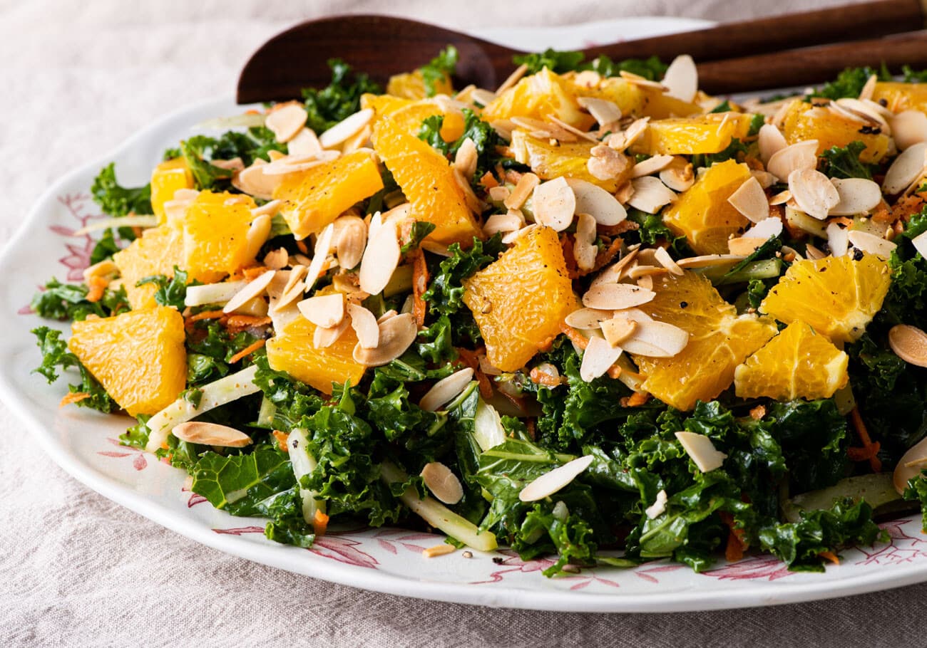 Kale fennel salad with oranges and sliced almonds on an oval platter with wooden spoons.