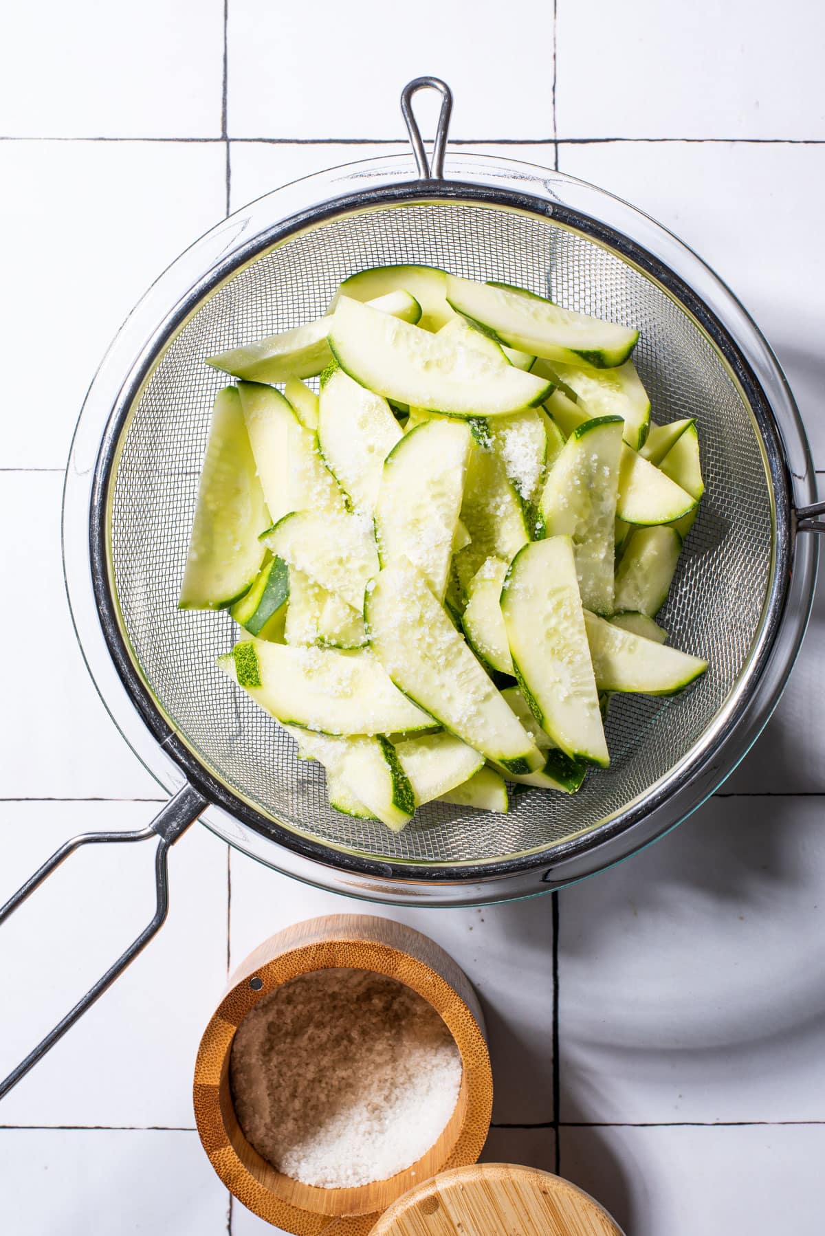 Sliced cucumbers in a mesh strainer next to salt.