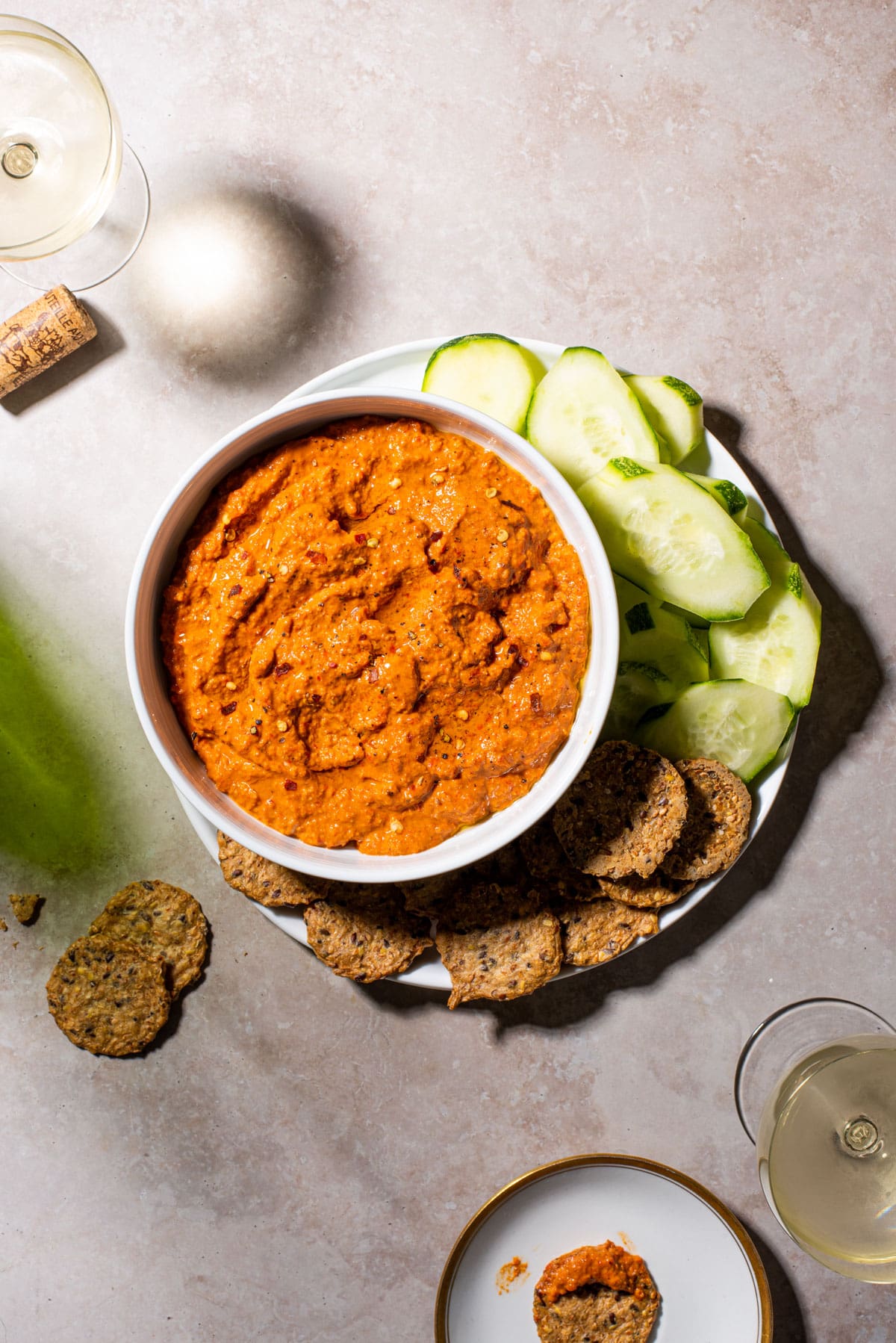 Muhammara dip in a white bowl next to cucumbers, crackers, and wine glasses.