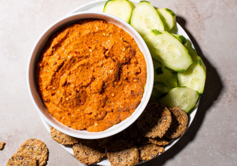 Muhammara recipe in a white bowl next to cucumber sliced and crackers.