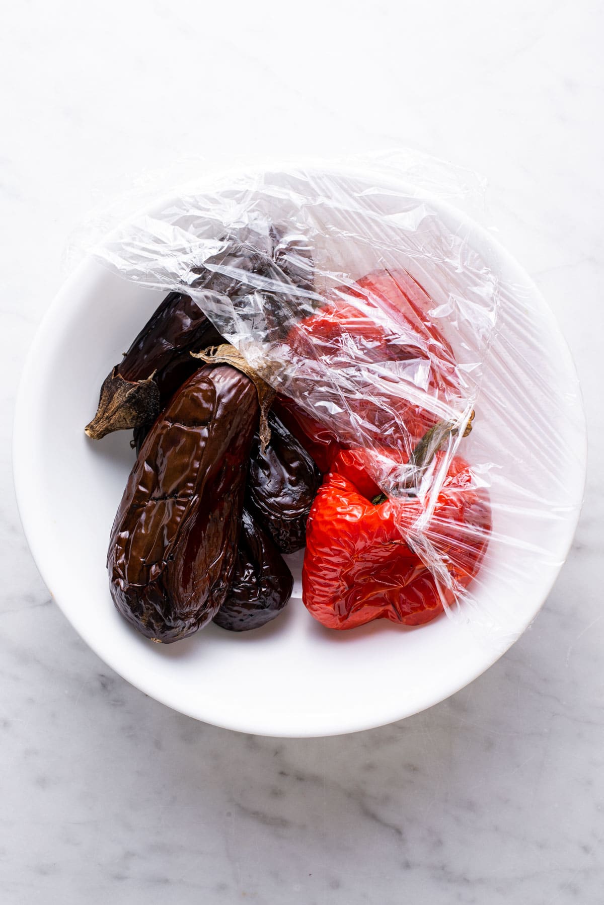 Roasted eggplants and peppers in a white bowl with plastic wrap.