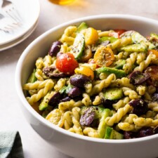Vegan pesto pasta salad with tomatoes, cucumbers, and olives in a beige bowl.
