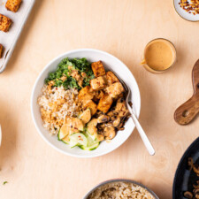 Assembling rice bowl with baked tofu, kale, cucumbers, mushrooms, and peanut sauce.