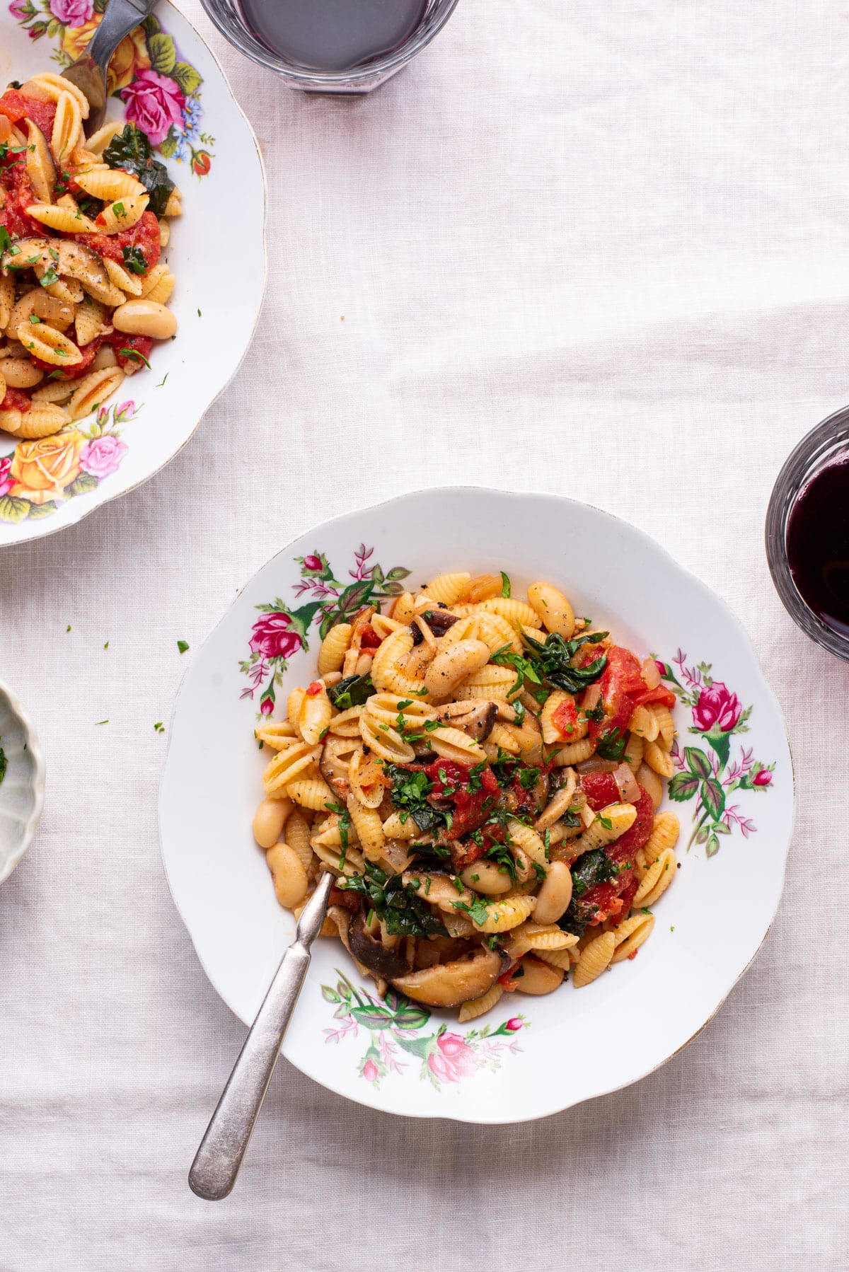 Two plates of pasta with beans and greens next to glasses of wine.