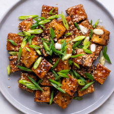 Pan-fried tofu cubes with sesame-garlic soy glaze, on a gray plate with sesame seeds and scallions.