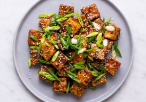 Pan-fried tofu cubes with sesame-garlic soy glaze, on a gray plate with sesame seeds and scallions.