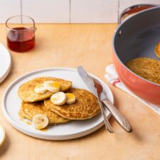 Vegan cornmeal pancakes on a white plate with bananas and maple syrup, next to pink Caraway sauté pan.