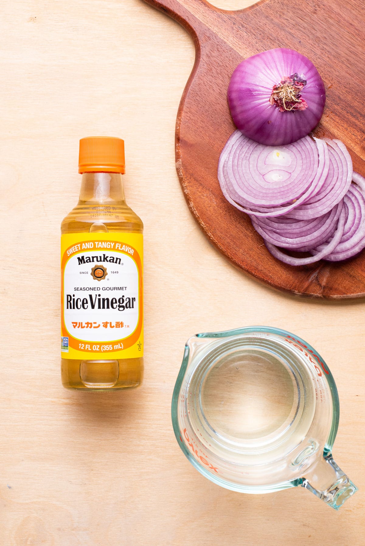 Seasoned rice vinegar, sliced red onion, and water, gathered on a wooden table.