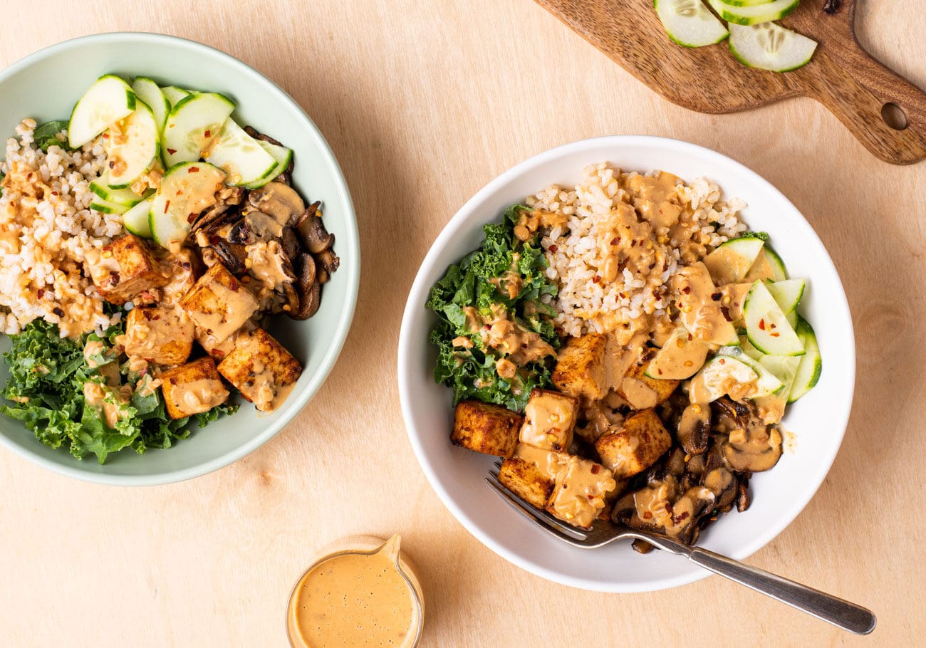 Two vegan grain bowls on a wooden table - with tofu, brown rice, cucumbers, peanut sauce.