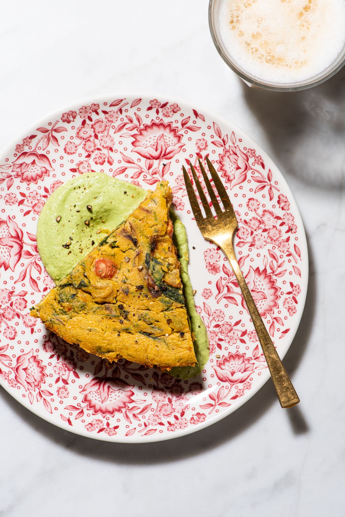 Slice of chickpea flour-based vegan frittata on a floral plate with cilantro cashew cream, next to a latte.