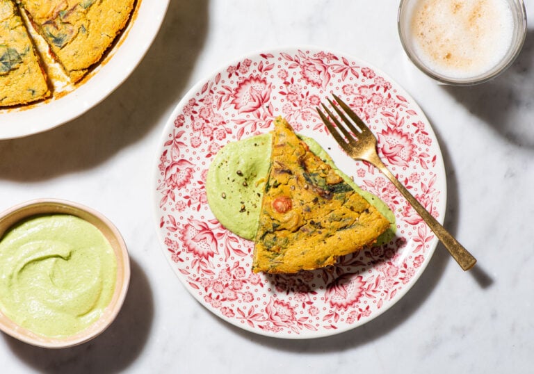 Slice of chickpea flour-based vegan frittata on a floral plate next to cilantro cashew cream and a latte.